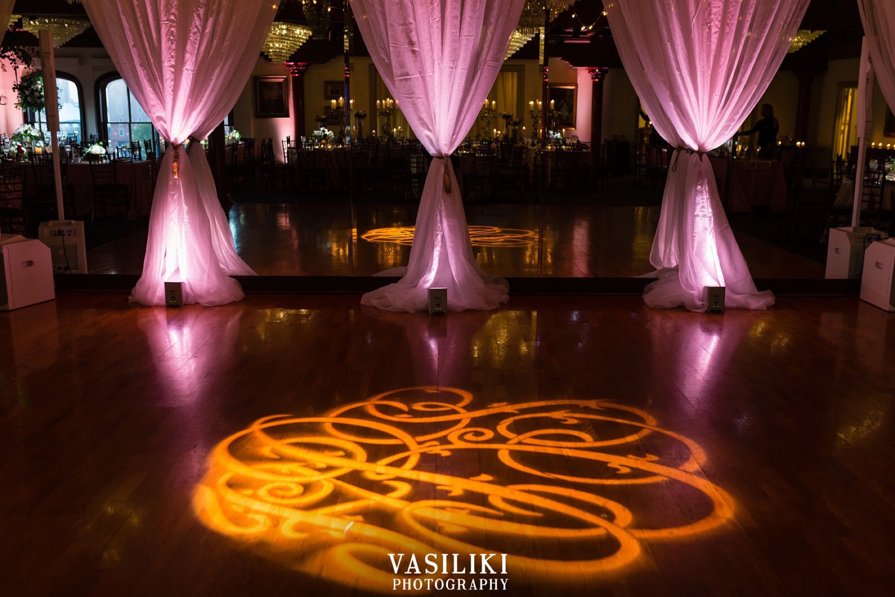A ballroom with draped uplighting and a gold monogram on the floor